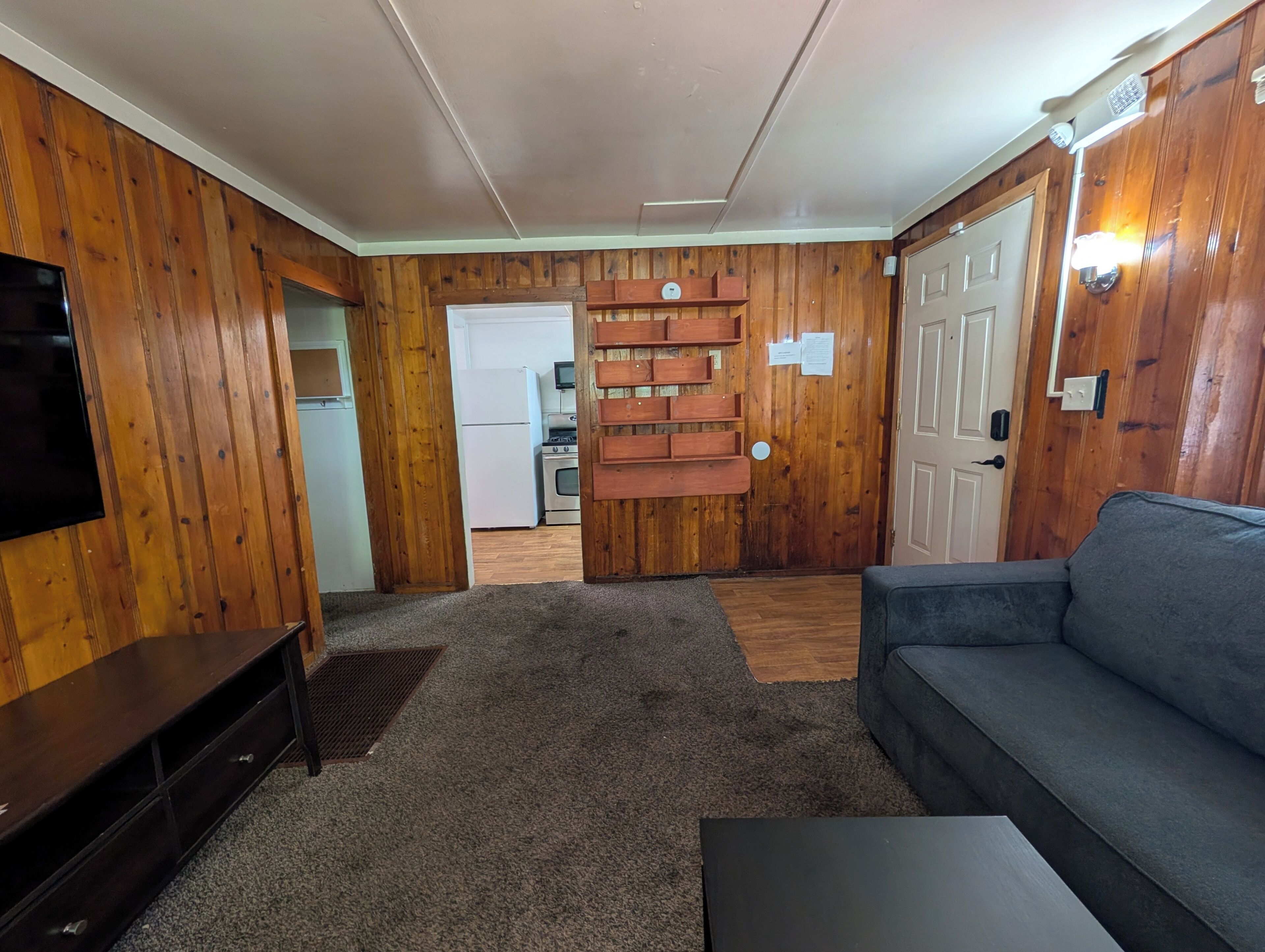 Un soggiorno con divano, televisione, parete in legno, cucina sullo sfondo e una porta.