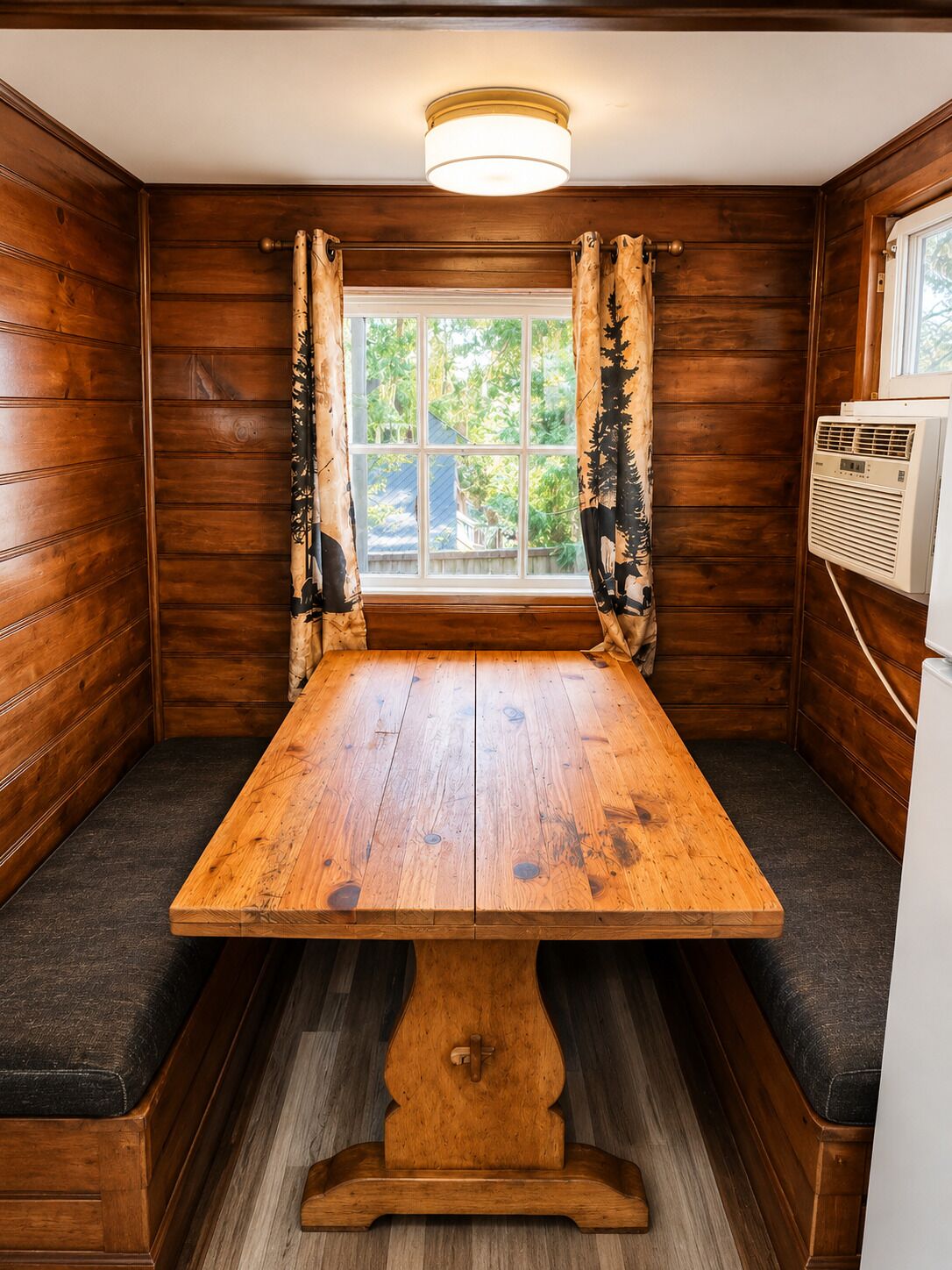 Un intérieur en bois avec une table centrale, des bancs rembourrés, une fenêtre avec des rideaux et un appareil de climatisation.