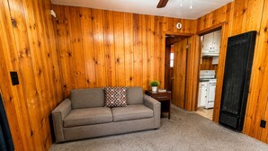 A room with a wooden paneled wall, a grey sofa with a patterned pillow, a small table with a plant, and a kitchen area visible through an open door. - Crest Lodge Historic Mountain Inn (Crestline)