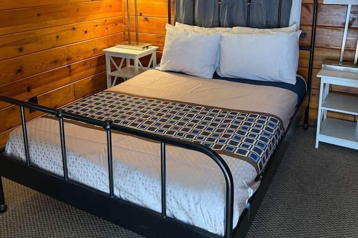 Un lit simple avec un cadre en métal, une tête de lit en bois et un rideau gris. Il y a une petite table de chevet avec une lampe et une chaise blanche.