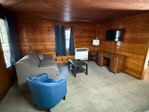 Ein gemütliches Zimmer im Chalet-Stil mit einer Couch, einem blauen Sessel, einem kleinen Tisch, einem Fernseher und einem Schreibtisch mit Schubladen.