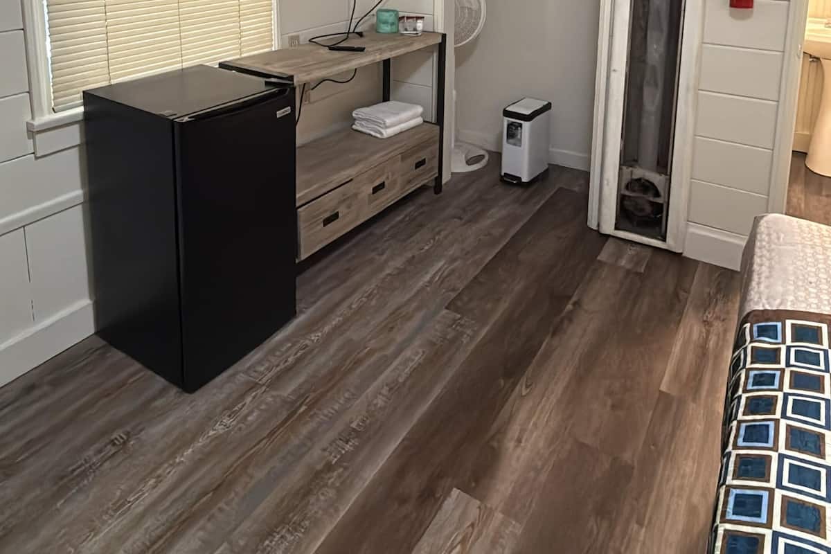 Une pièce de taille modeste, avec un plancher en bois, un réfrigérateur noir, un téléviseur fixé au mur, un petit ventilateur et un extincteur.