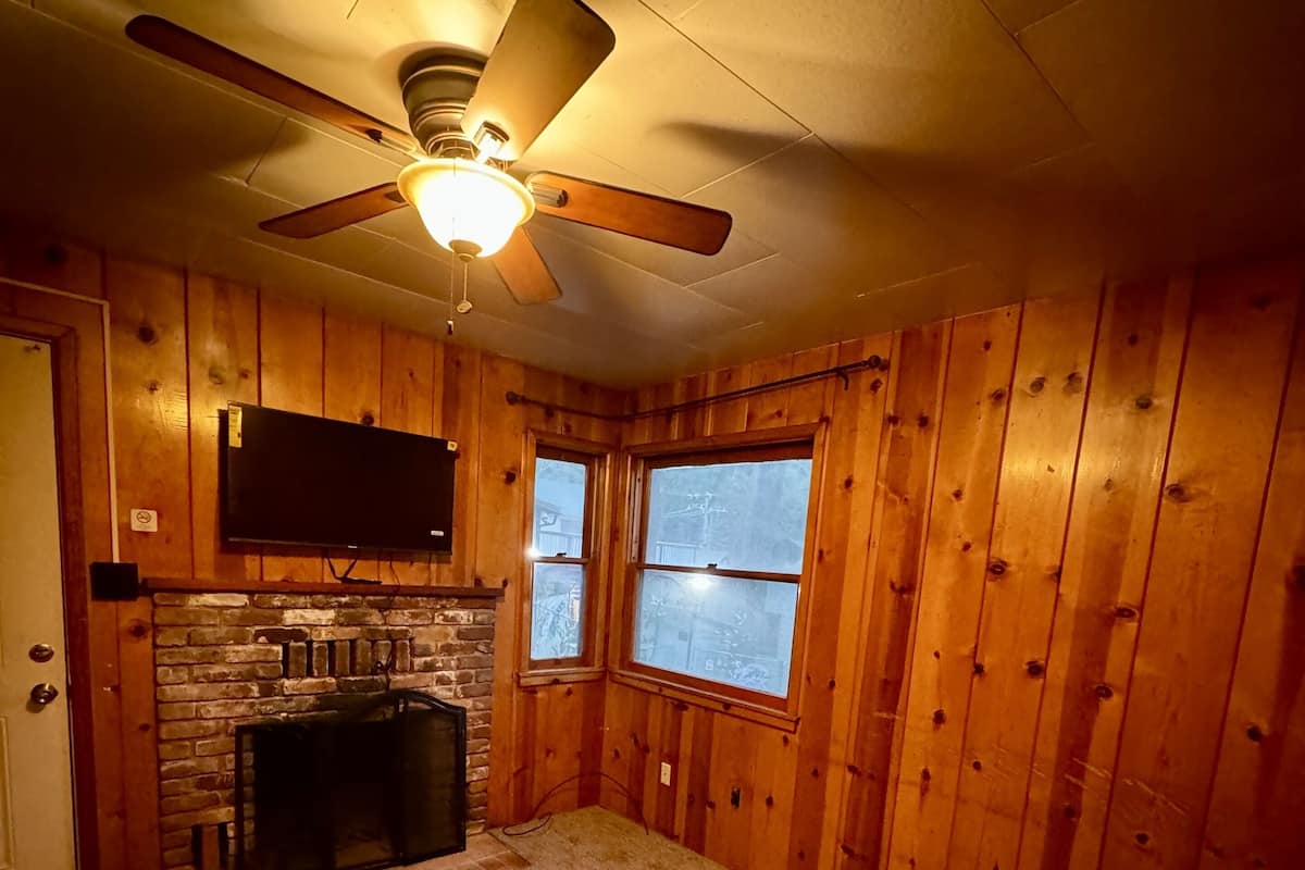 Une pièce chaleureuse avec des murs en bois, une cheminée en pierre, un ventilateur de plafond et une grande fenêtre.