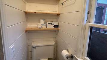 A small, compact bathroom with a toilet, a wooden shelf above the toilet, and a window to the right.