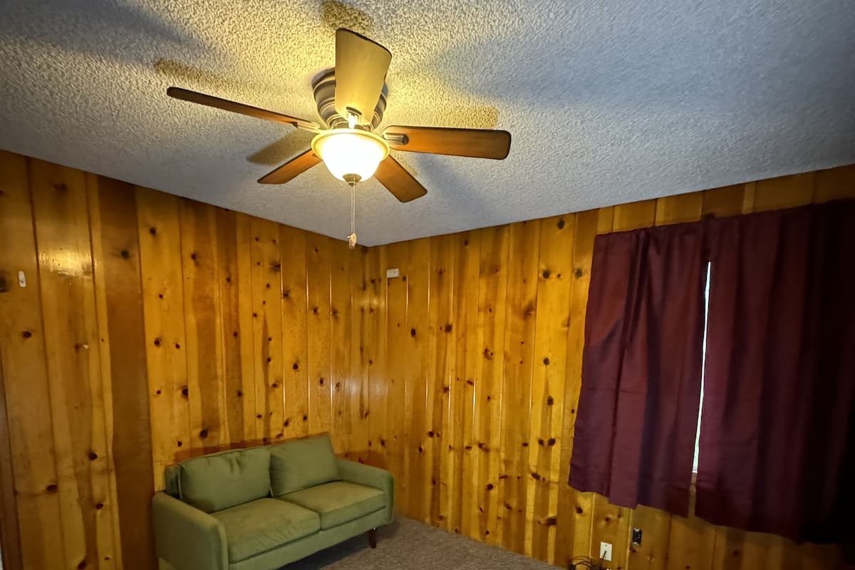Une pièce aux murs recouverts de lambris en bois, avec un canapé vert, un ventilateur de plafond et une fenêtre avec des rideaux.