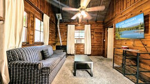 A cabin interior with a checkered sofa, a coffee table, a ceiling fan, and a television displaying Lake Gregory. - Crest Lodge Historic Mountain Inn (Crestline)