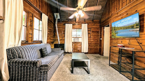 L’intérieur d’une cabane est aménagé avec un canapé à carreaux, une table basse, un ventilateur de plafond et un téléviseur qui diffuse des images du lac Gregory.