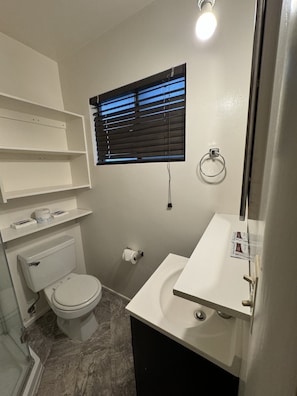 A bathroom with a toilet, sink, and window with blinds. There are shelves above the toilet and a towel rack on the wall. - Crest Lodge Historic Mountain Inn (Crestline)