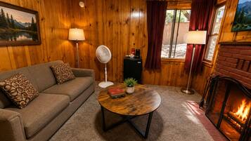 A cozy living room with a wooden interior, a fireplace, a sofa with patterned cushions, a coffee table with books, and a standing fan.