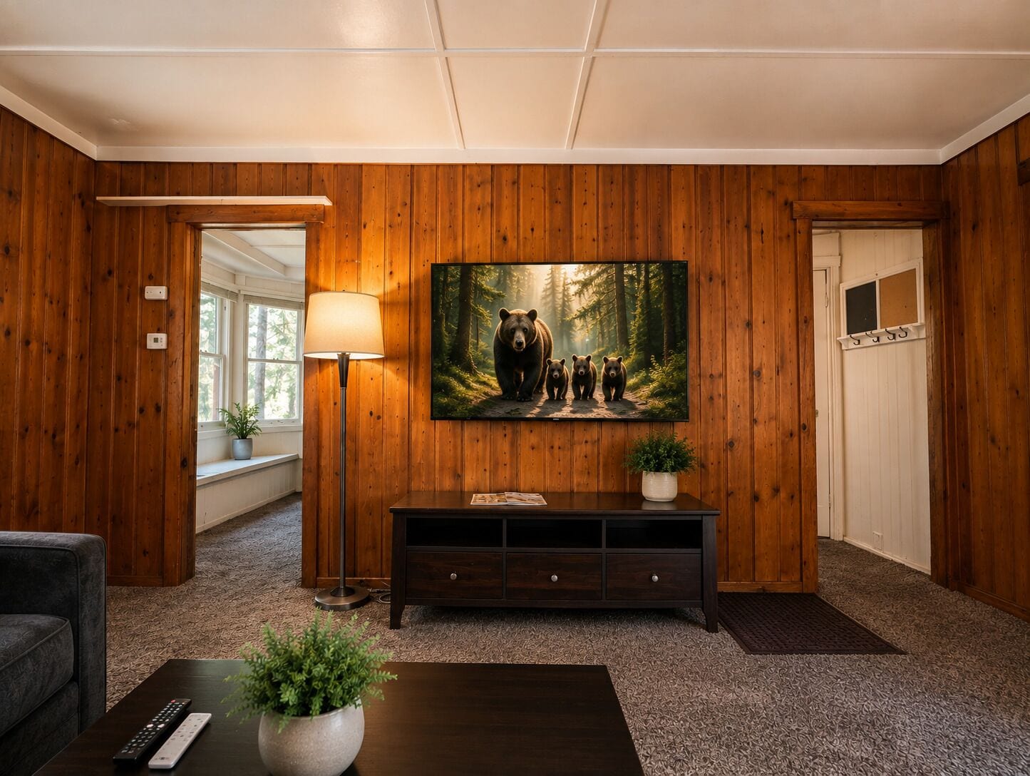 Ein Zimmer mit Holzvertäfelung, einem großen Gemälde von Bären, einer Stehlampe, einem dunklen Konsolentisch und einer Fernbedienung auf einem Couchtisch.