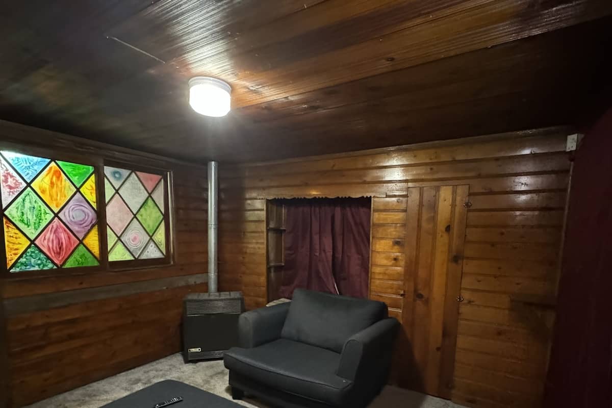 Une pièce chaleureuse aux murs en bois, avec un canapé noir, un pouf assorti et une fenêtre colorée en vitrail.