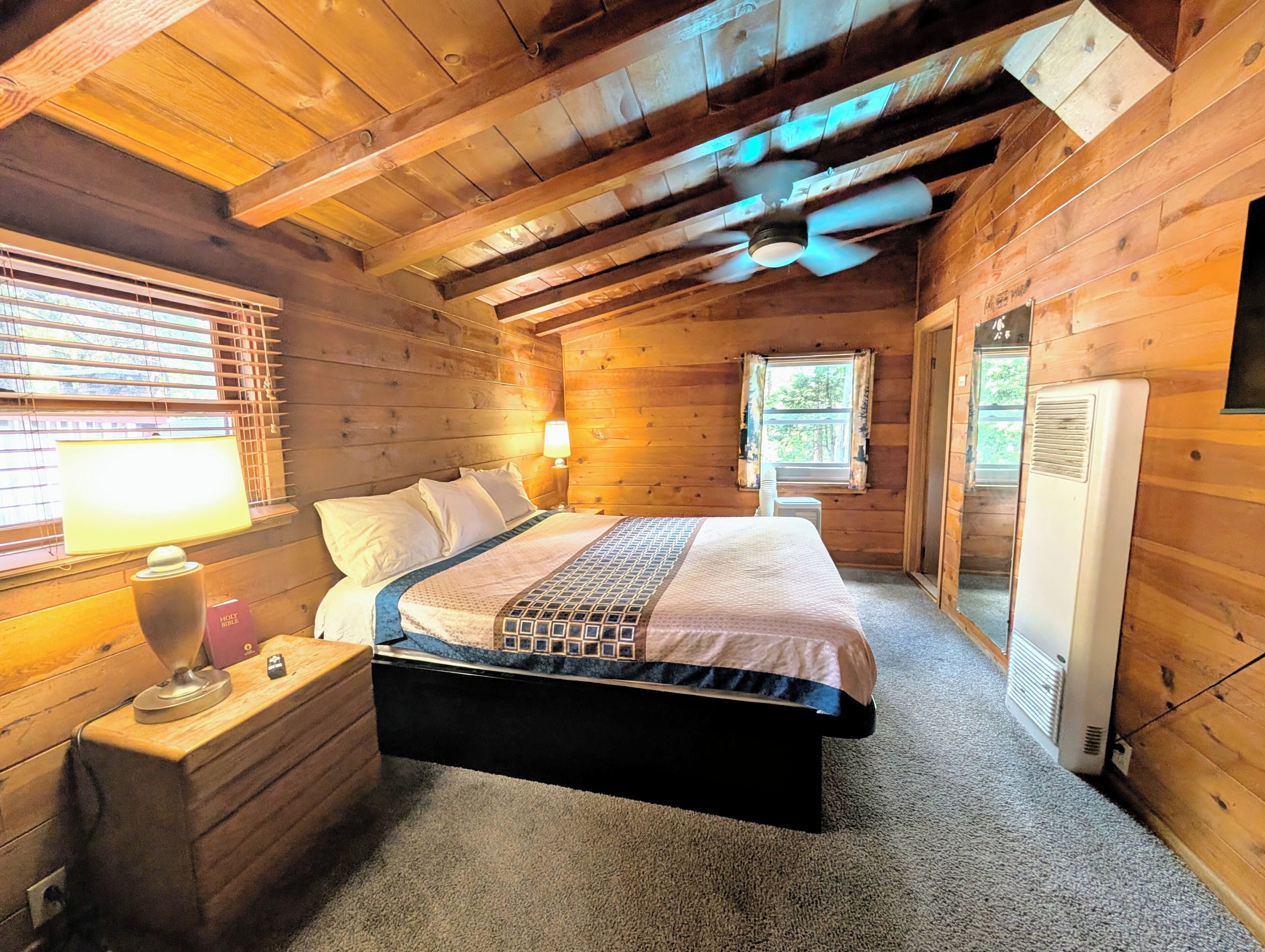 A wooden cabin bedroom with a bed, bedside lamp, and a window with blinds.