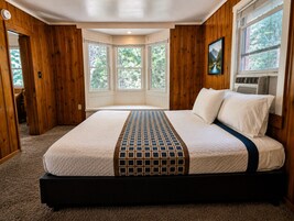 Un dormitorio con una cama grande, paredes de madera, una ventana con vistas a árboles y un equipo de aire acondicionado.