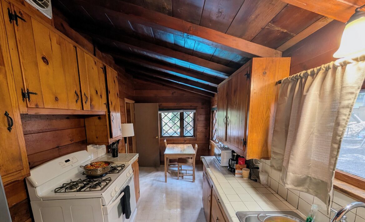 A wooden cabin interior with a kitchen area featuring a stove, a table, and a window.