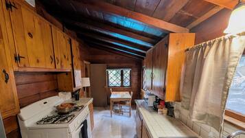 A wooden cabin interior with a kitchen area featuring a stove, a table, and a window.