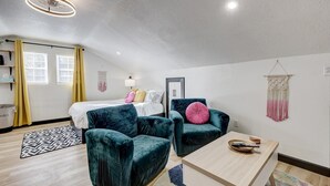 A modern, well-lit room with a bed, two teal armchairs, a wooden coffee table, and a macrame wall hanging. - The Crow's Nest:  Executive Loft (Webb City)