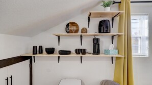 A kitchen with wooden shelves holding cups, plates, and a coffee maker. There is a trash bin and a chair in the room. - The Crow's Nest:  Executive Loft (Webb City)