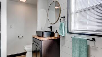 A modern bathroom with a wooden floor, a black sink, a mirror, and a window with blinds.