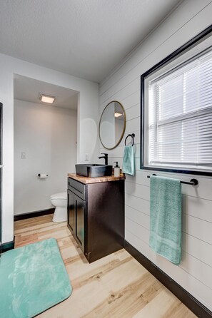A modern bathroom with a wooden floor, a black sink, a mirror, and a window with blinds. - The Robin's Egg: Downstairs Studio (Webb City)
