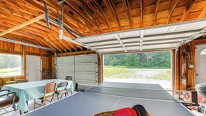 A ping pong table in a wooden room with a garage door, chairs, and a basket of ping pong paddles. - Fire Pit & On-Site Pond: Wooded Walton Home! (Walton)