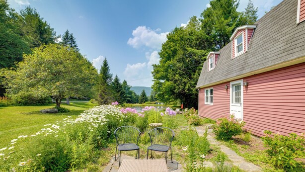 A red house with a garden patio featuring a table and chairs, surrounded by lush greenery and flowers. - Fire Pit & On-Site Pond: Wooded Walton Home! (Walton)