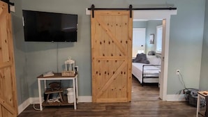 A room with a sliding barn door, a mounted TV, a small table with a 'home' sign, and a bed in the background. - Pickwick Getaway in the Groves 7th NT Free!
(Counce)