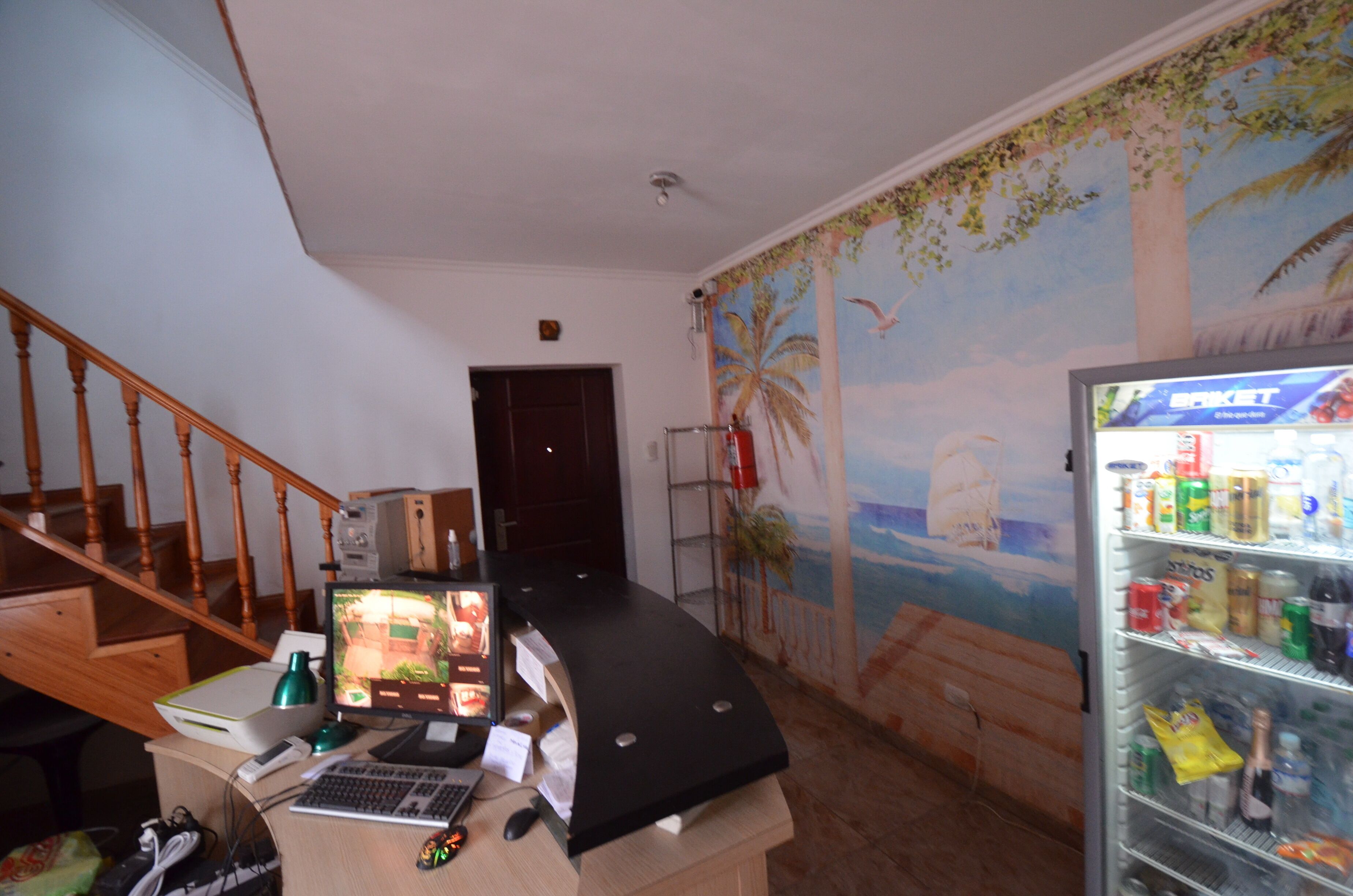Una habitación con una escalera de madera, un ordenador de escritorio, un refrigerador lleno de bebidas y un mural con una playa.