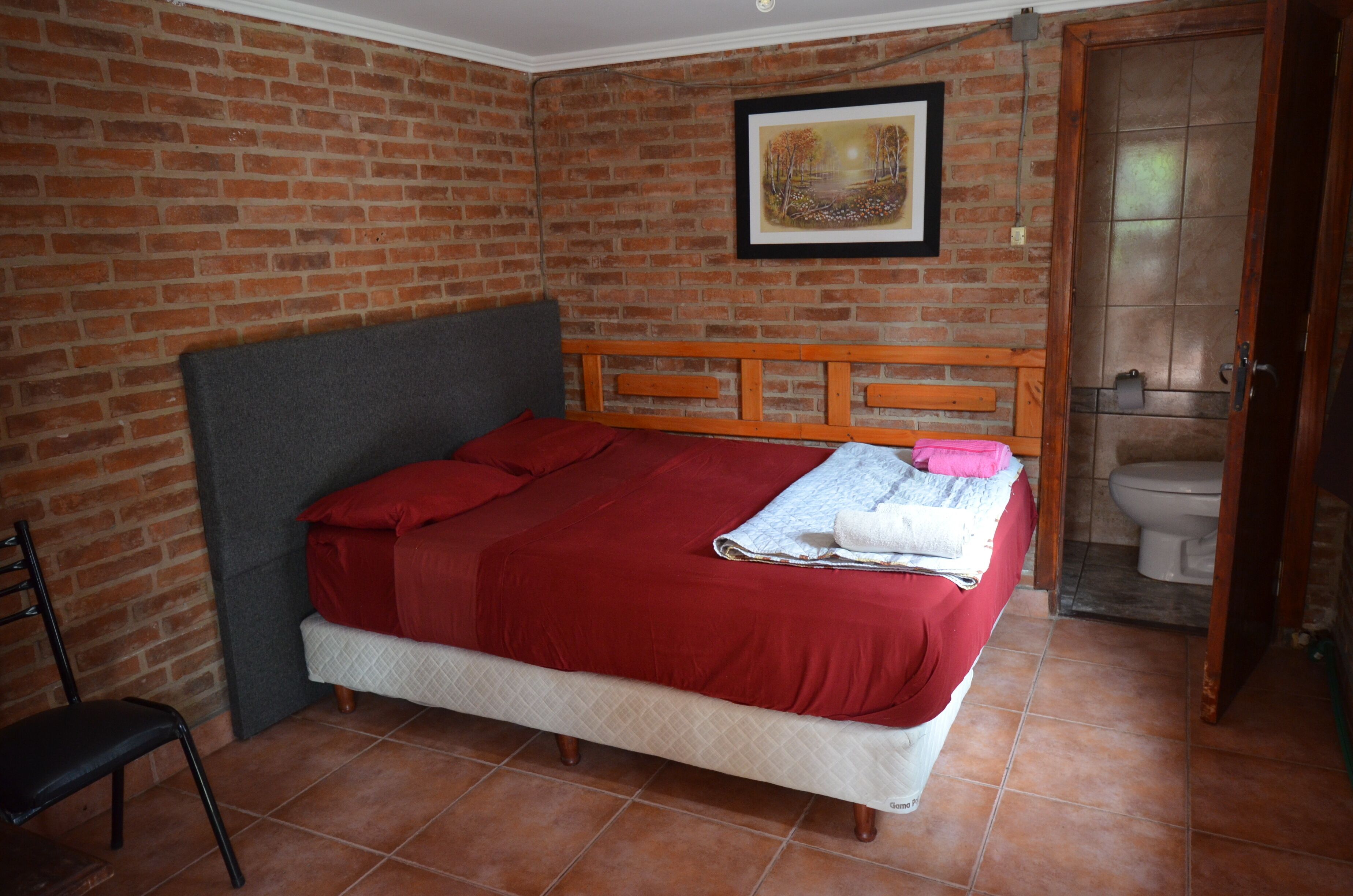 Un dormitorio con pared de ladrillo, cama con cubrecama rojo, cabecera de madera, un cuadro enmarcado en la pared, y un inodoro visible en la habitación contigua.