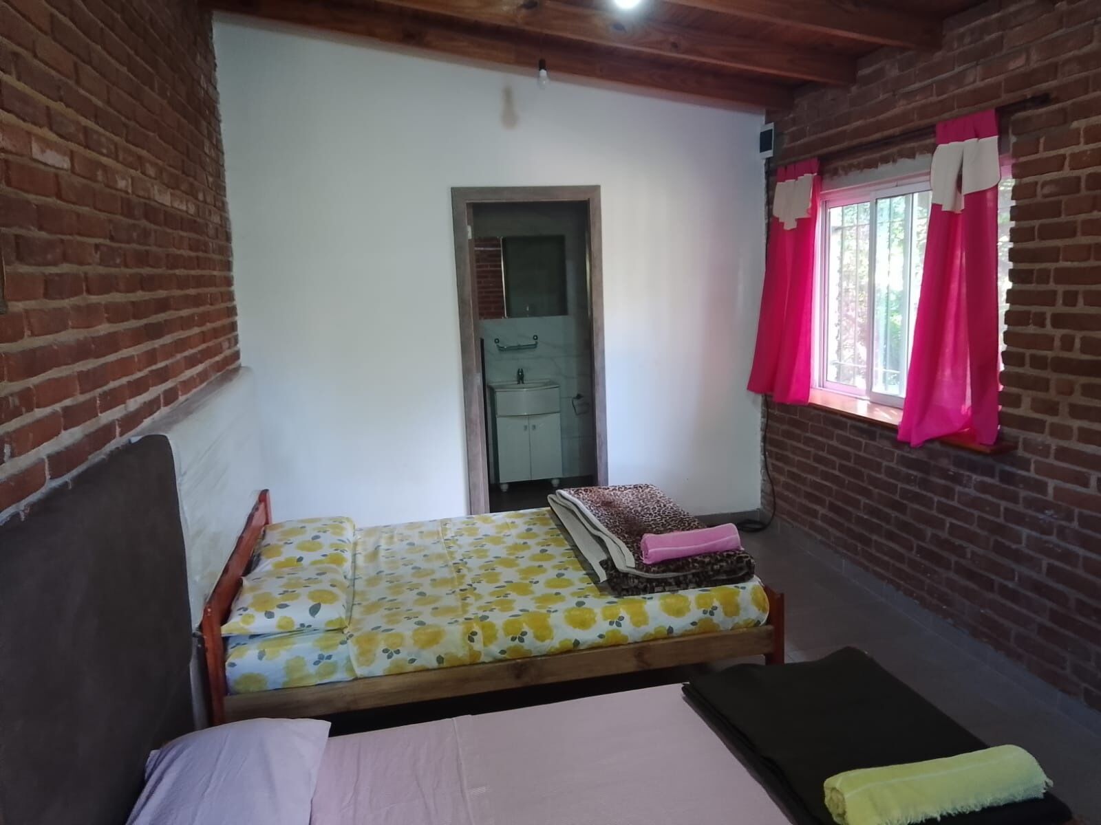 Une petite pièce accueillante, avec un lit simple, une tête de lit en bois, une fenêtre ornée de rideaux roses et un mur en briques.