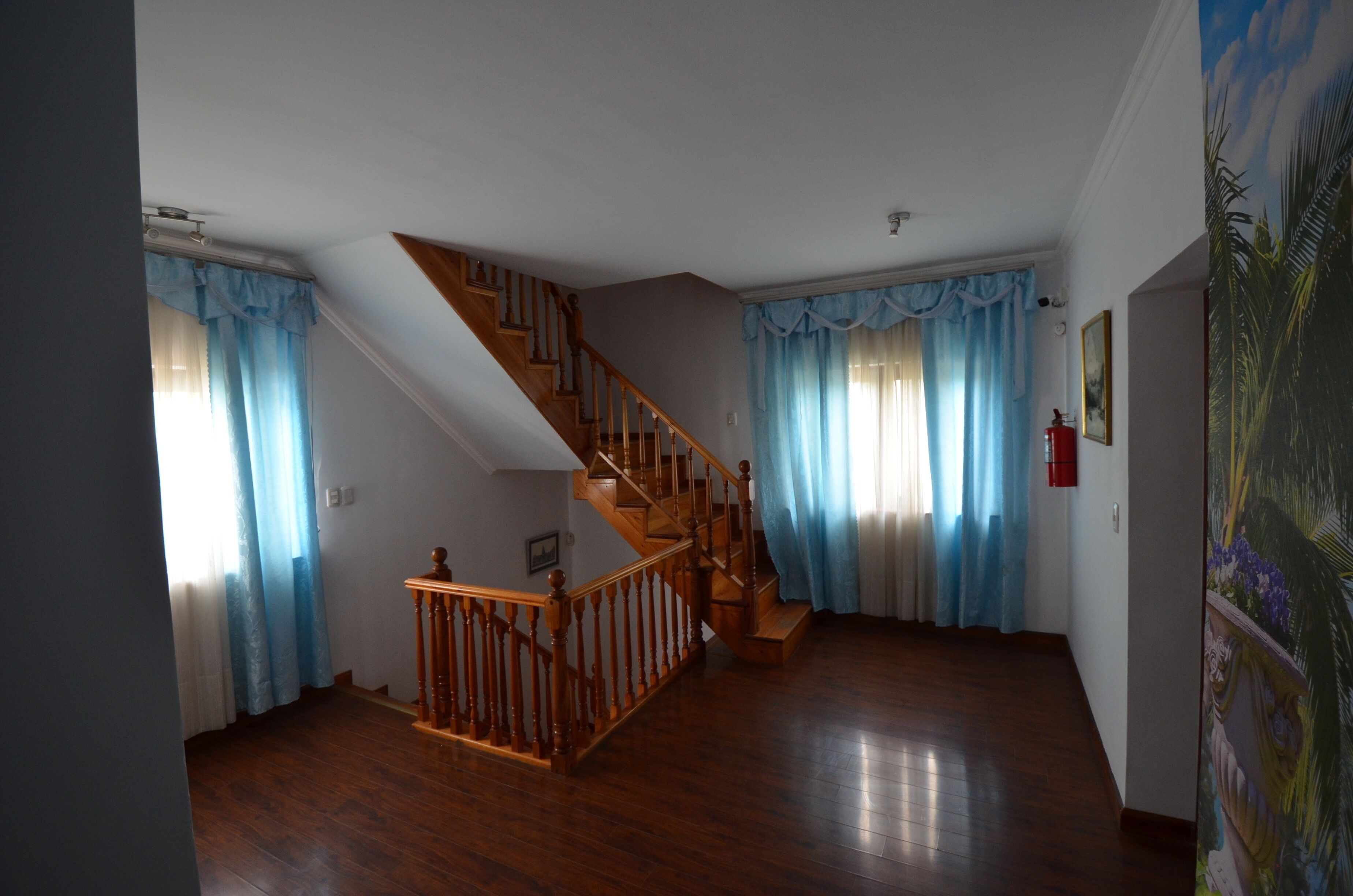 A spacious room with a wooden staircase, a large window with blue curtains, and a painting on the wall.