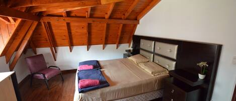 A bedroom with a wooden ceiling, a bed with a headboard, a nightstand, a chair, and a small table with a plant.