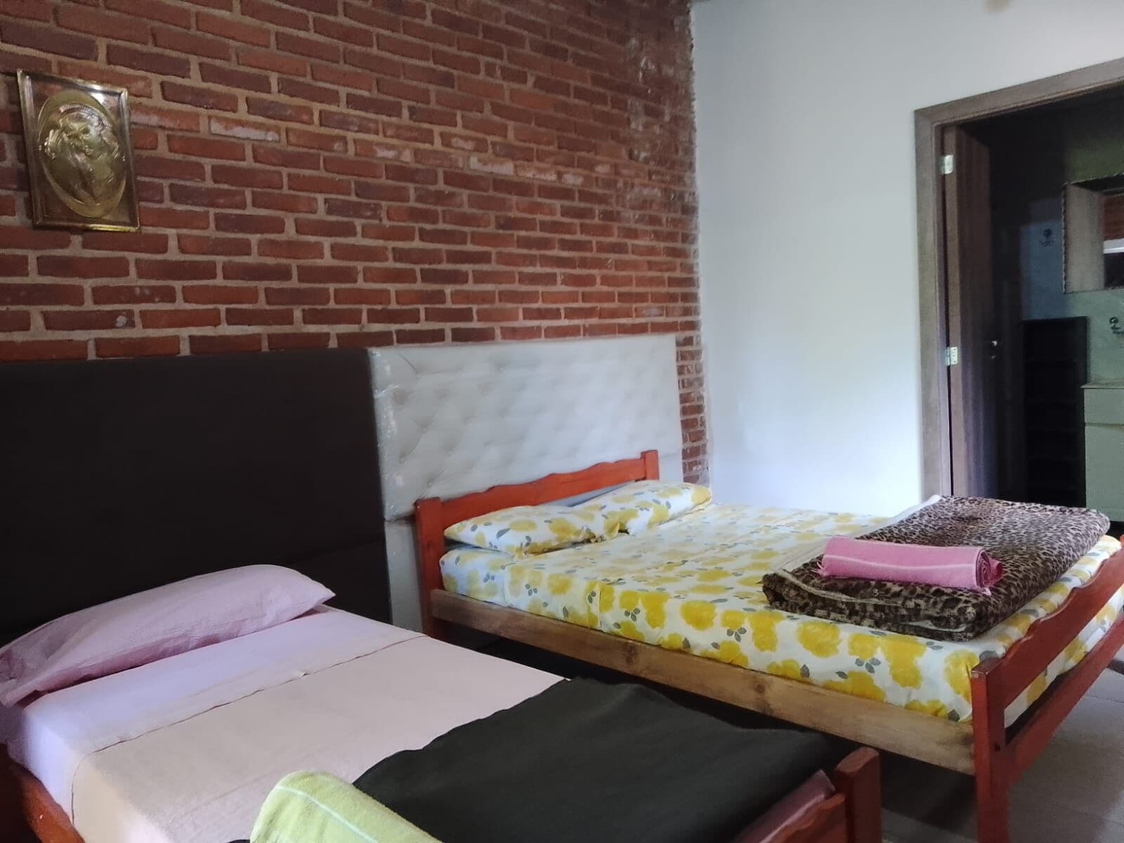 Quarto com parede de tijolos, cama com colcha floral, cabeceira de madeira e uma pequena mesa com toalha rosa.