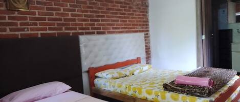 Quarto com parede de tijolos, cama com colcha floral, cabeceira de madeira e uma pequena mesa com toalha rosa.