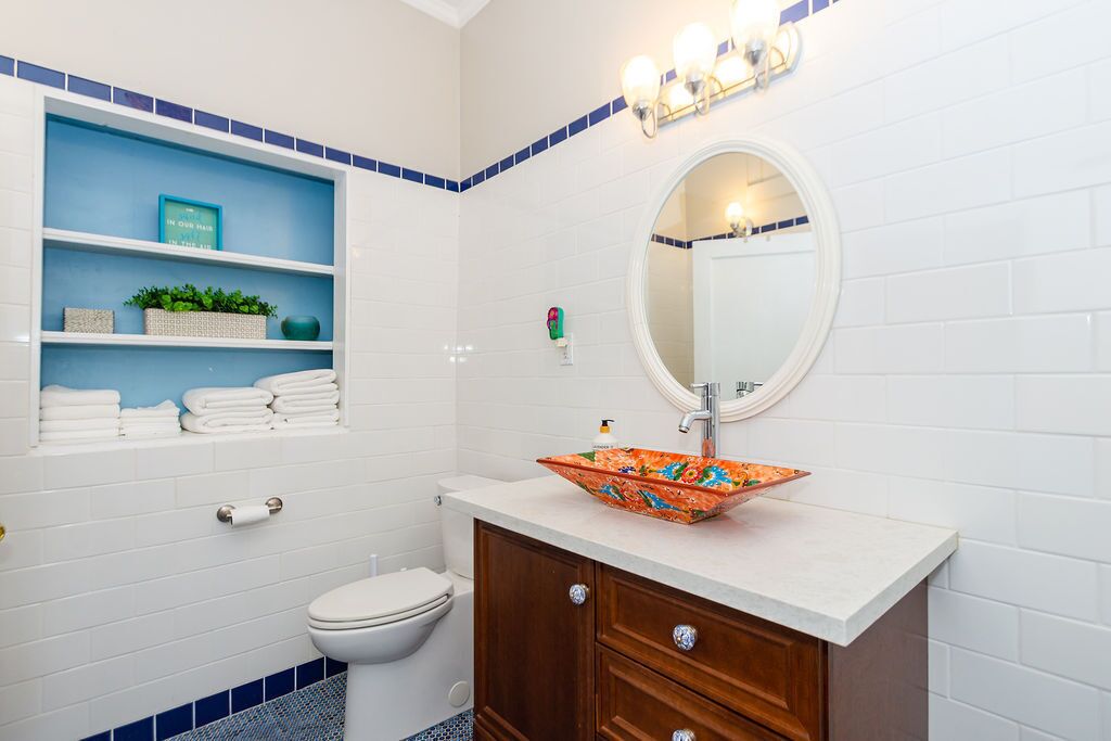 Ein Badezimmer mit einer weißen Toilette, einem hölzernen Waschtisch mit einem dekorativen Becken sowie einem blau gefliesten Ablagebereich mit Handtüchern und Pflegeprodukten.