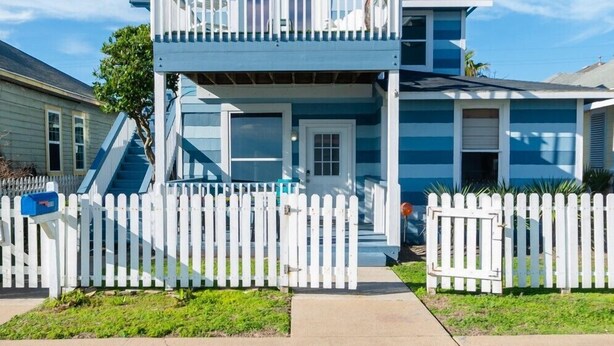 Une maison à deux étages avec un balcon, une clôture blanche en bois et un ciel bleu dégagé.
