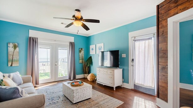 A living room with teal walls, a ceiling fan, a TV, a white dresser, and a large window. - Upper Unit Beach Rental in Galveston Near Strand, Pleasure Pier & Restaurants! (Galveston)