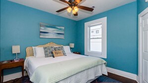 A bedroom with a bed, a ceiling fan, a nightstand, a lamp, a painting, and a window. - Upper Unit Beach Rental in Galveston Near Strand, Pleasure Pier & Restaurants! (Galveston)