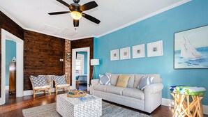 A living room with a blue wall, wooden accents, a ceiling fan, a grey sofa, wicker chairs, a patterned ottoman, and framed pictures. - Upper Unit Beach Rental in Galveston Near Strand, Pleasure Pier & Restaurants! (Galveston)
