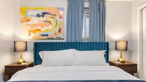 Un lit bien fait, recouvert de draps blancs, flanqué de deux tables de chevet avec des lampes, dans une pièce dotée d’une tête de lit bleue et d’un tableau abstrait coloré accroché au mur.