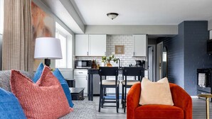 A modern living room with a grey sofa, red and blue cushions, and a red armchair. A kitchen with white cabinets and a black countertop is visible in the background. - Westlake Elegance Unit 3 (Westlake)