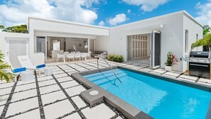 Een modern buitenzwembad met een helderblauwe lucht, een barbecuegrill en een terras met zitplaatsen.