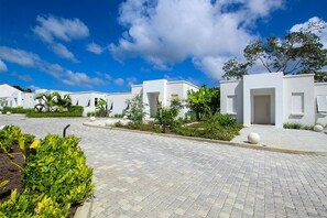 A row of white buildings with a paved walkway, surrounded by greenery and a clear sky.
