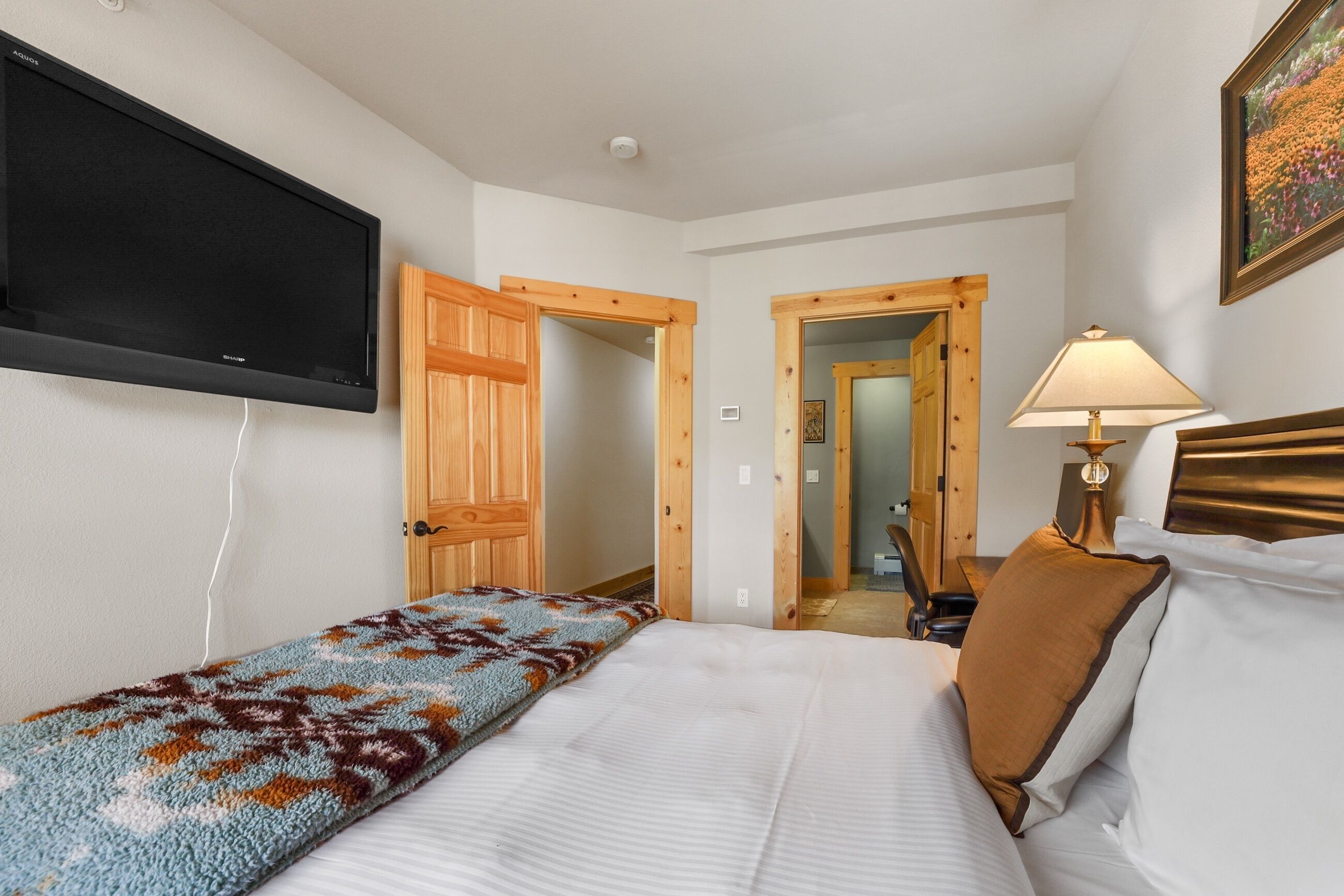A hotel room with a bed, a TV mounted on the wall, a wooden door, a lamp, and a painting on the wall.
