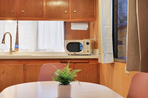 A compact kitchen with wooden cabinets, a white countertop, a vintage microwave, a sink with a gold faucet, and a small potted plant on the table.