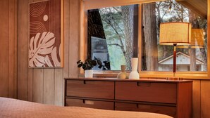 A bedroom with a wooden dresser, a bed with a textured blanket, a window with a view of trees, and a wall with a leaf-patterned artwork. - Modern Cabin (Walnut) (Dunsmuir)