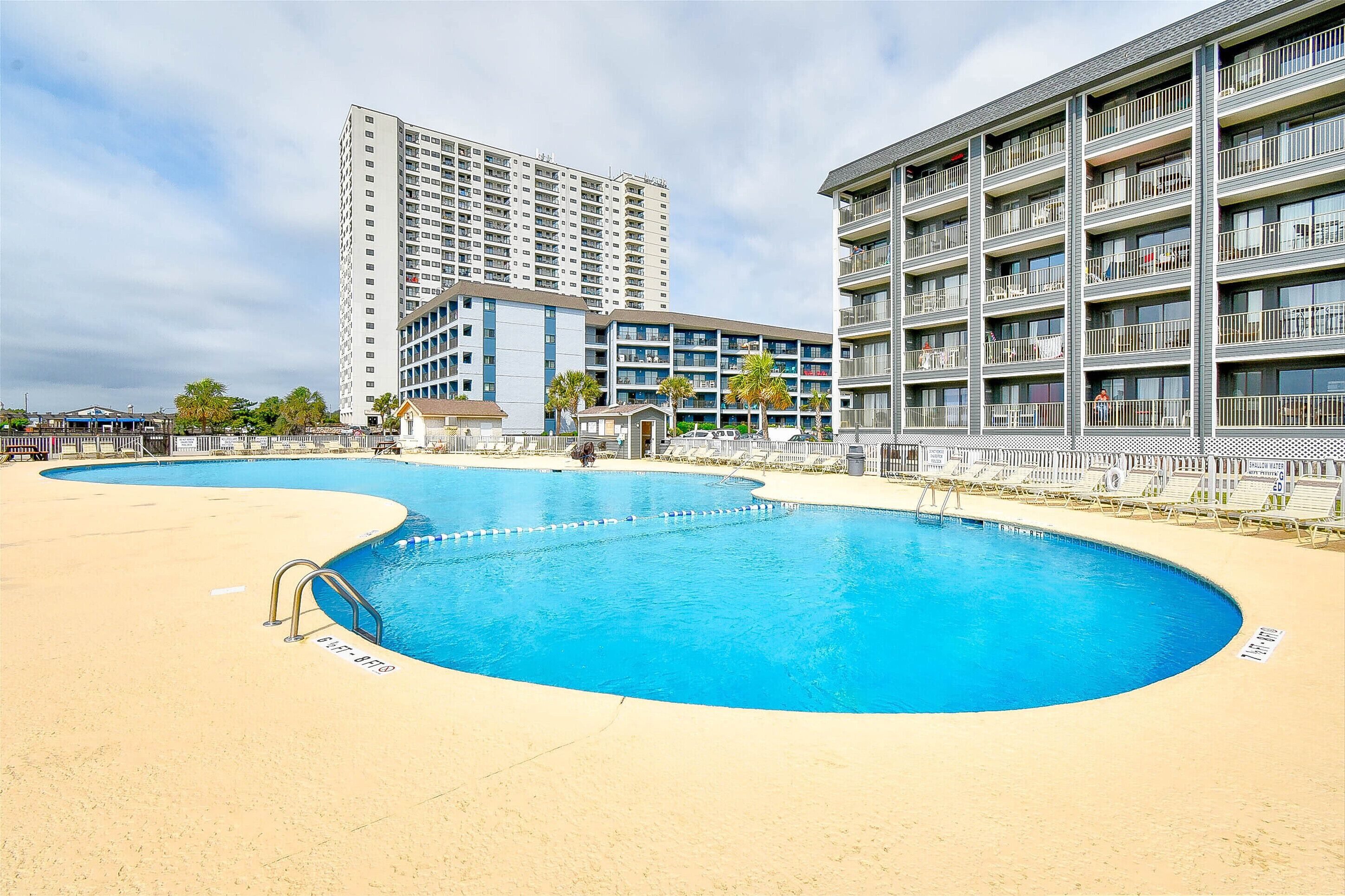 Una piscina de diseño curvo, rodeada por una zona arenosa. En el fondo, edificios de varios pisos con balcones.