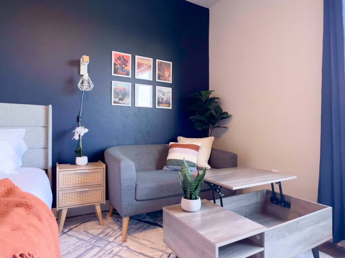 Ein modernes Hotelzimmer mit Bett, grauem Sofa, Nachttisch aus Holz und einem kleinen Tisch mit Pflanze.