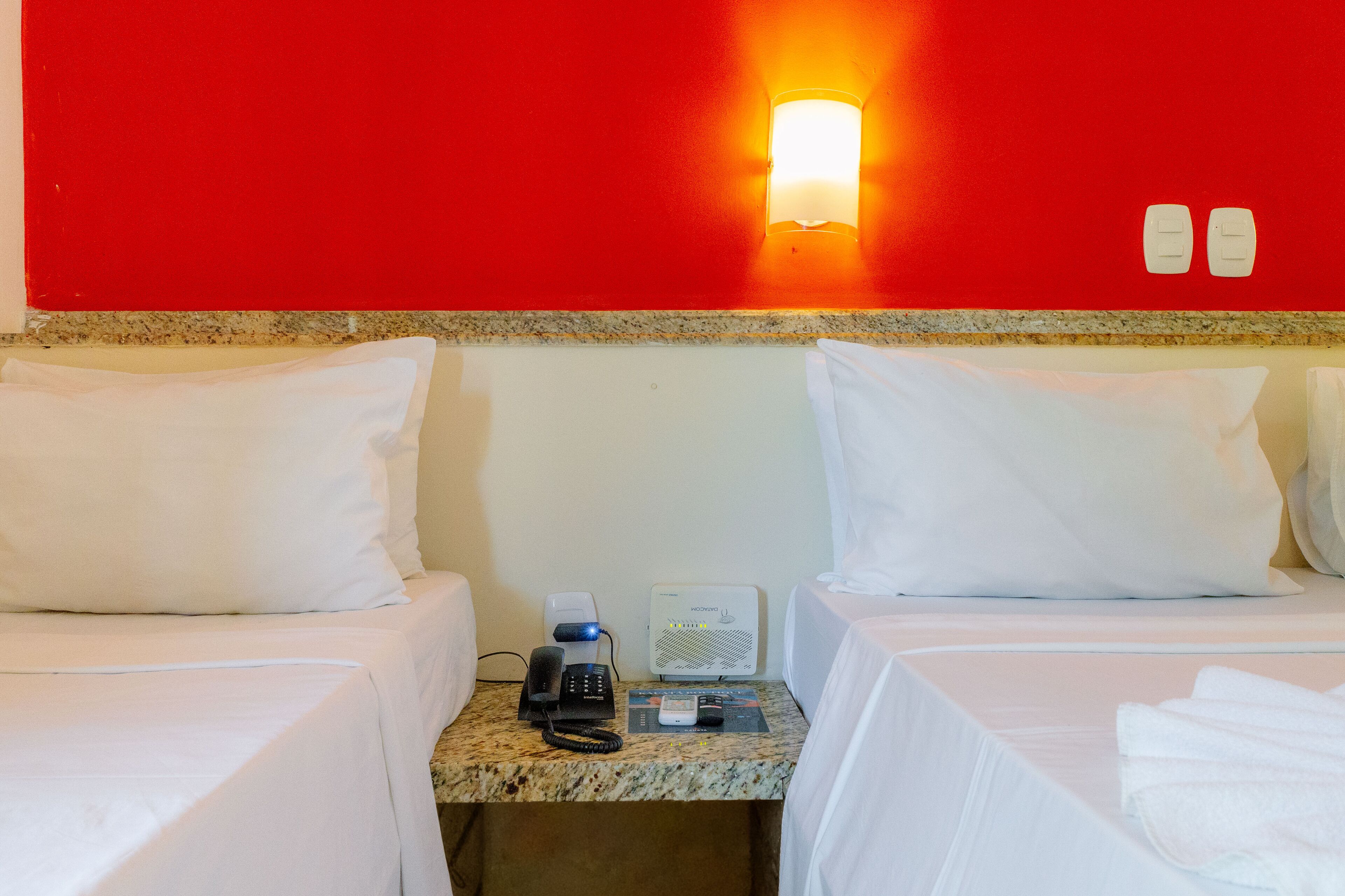 A hotel room with two beds, a nightstand with a phone and a thermostat, and a red wall with a light fixture.