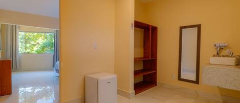 Una habitación de hotel con refrigerador compacto, armario de madera, piso de baldosas de mármol y baño con lavamanos y espejo.