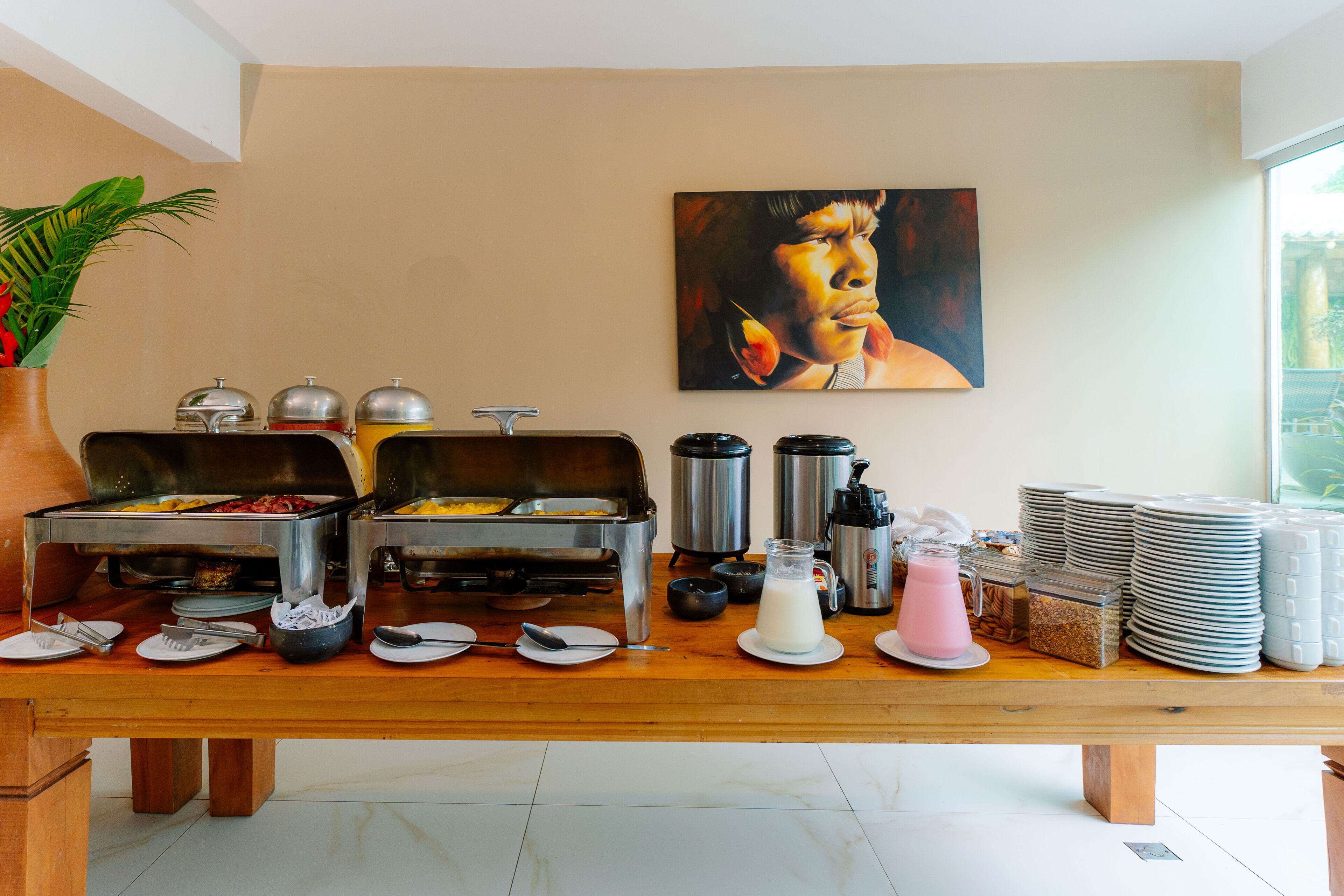 A buffet setup with various food items and beverages, a painting on the wall, and a potted plant.